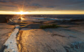 Auringonlasku Kylmäpihlajan majakkasaarella. Aallot lyövät sileään kallioon.