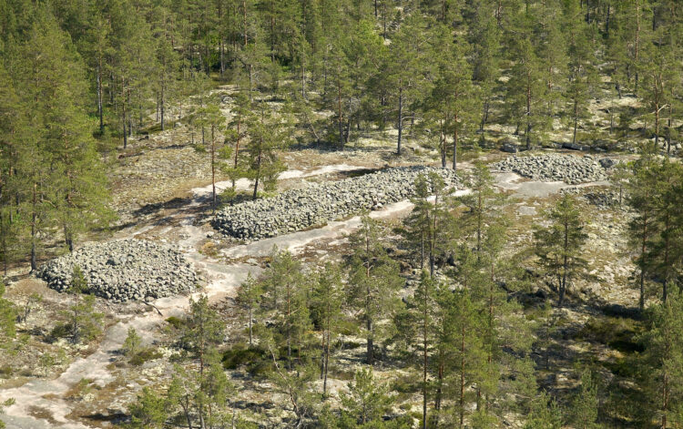 Aerial view of the Bronze Age burial site of Sammallahdenmäki. The picture shows a large, elongated stone tumulus in the centre and part of another tumulus to the left. The terrain is rocky forest.