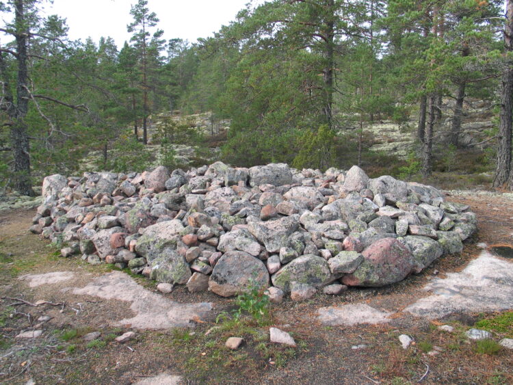 Photo of the Bronze Age burial mound area at Sammallahdenmäki. The picture shows the whole of one low stone cairn. The landscape in the picture is a rocky forest landscape. Coniferous trees can be seen in the background.