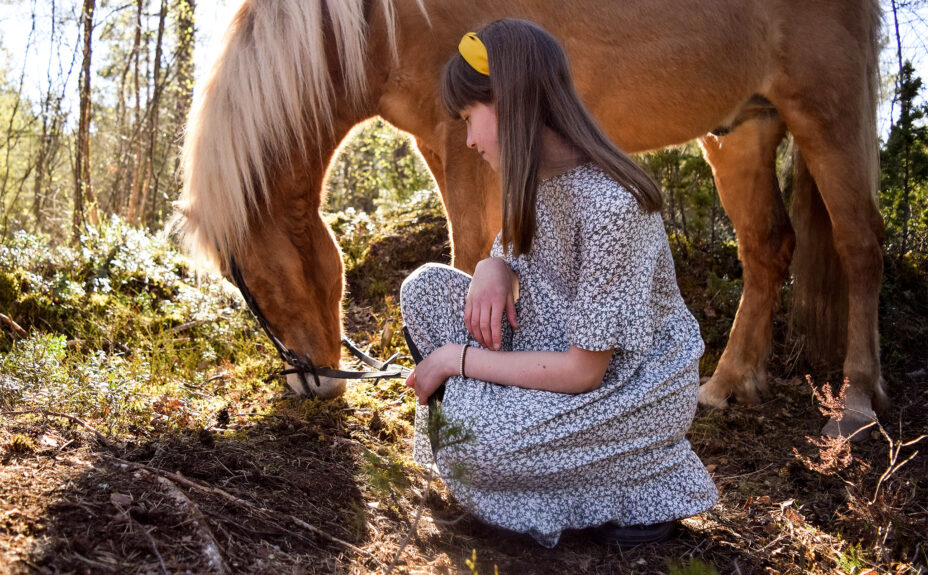 An Icelandic horse is eating in the background. A young woman in front of a horse.